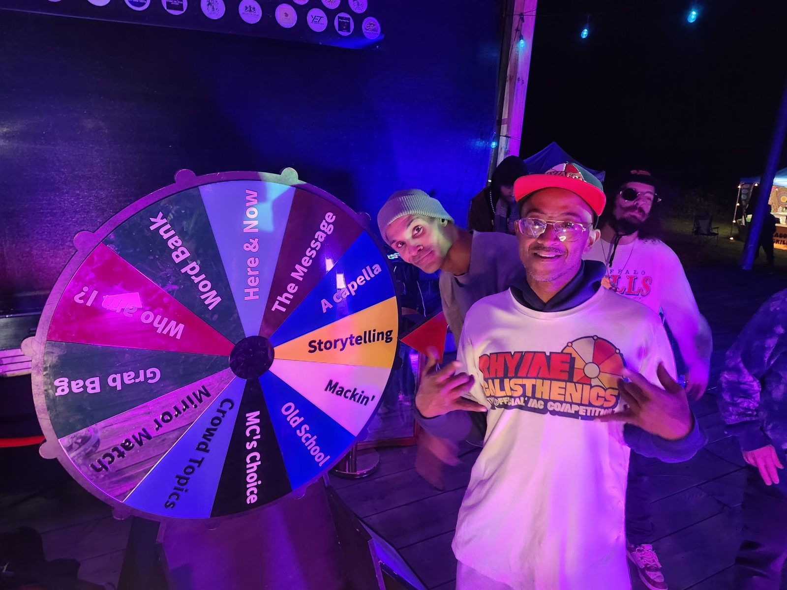 A spinning prompt wheel lit up at night beside two artists