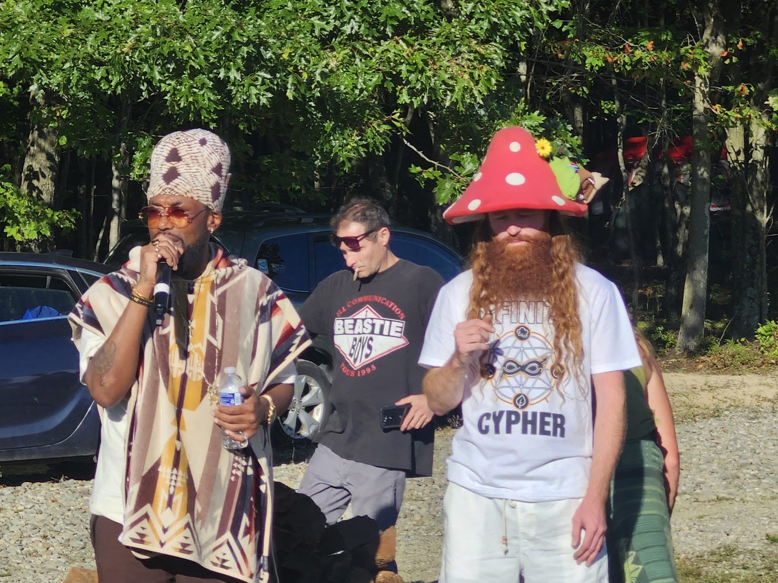Two artists in hats, one on the mic, trees behind them